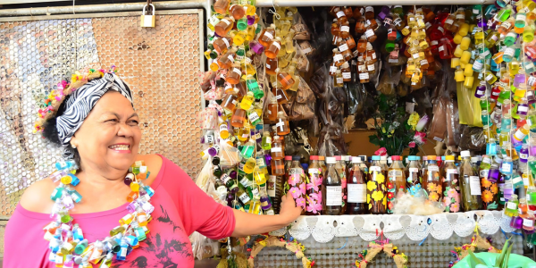 Dona Onete sorrindo, de lenço na cabeça e colar colorido, diante de uma banca de garrafas de essências