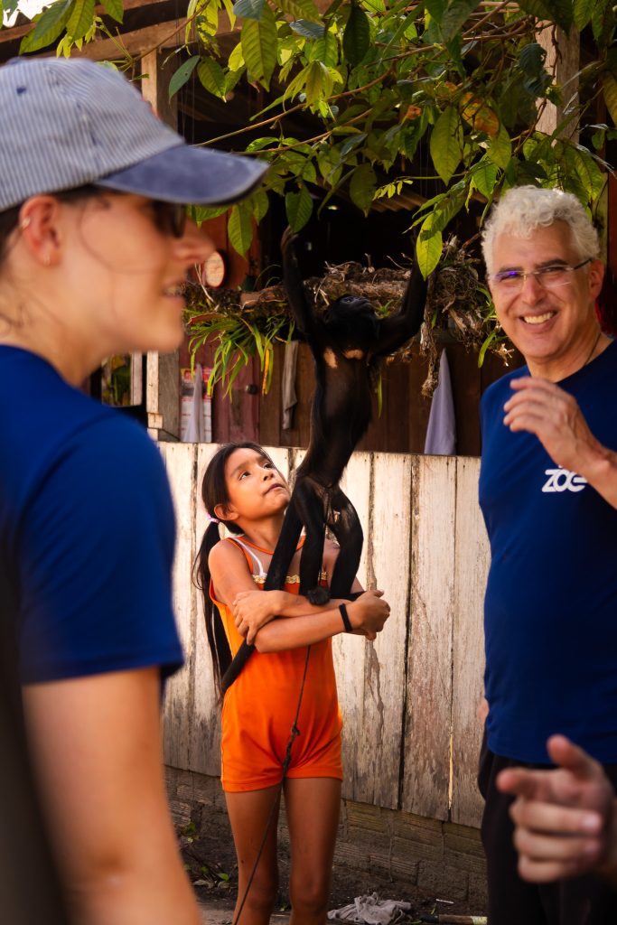 Dois voluntários da ONG Zoé, de camiseta azul, conversam com uma menina que segura um macaco no colo, em uma comunidade ribeirinha da Amazônia