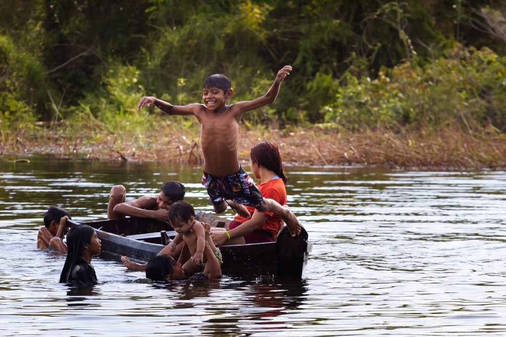 Crianças brincam em um rio da Amazônia: um menino salta de uma canoa de braços abertos enquanto outras nadam ao redor