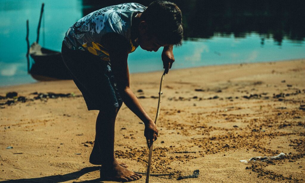 Menino descalço desenha na areia com um graveto na beira do rio Arapiuns, com uma canoa ao fundo